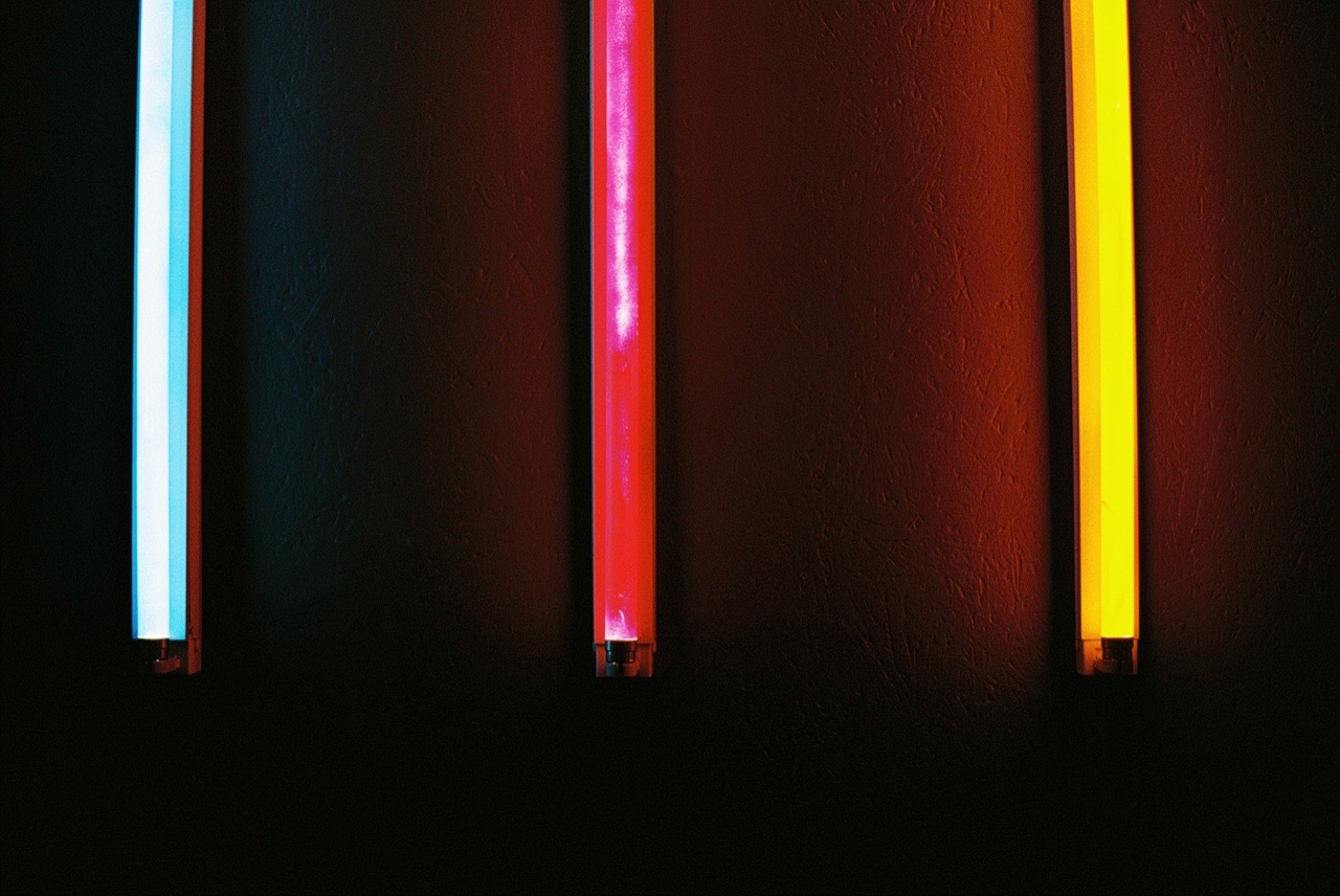 Drei leuchtende Neonröhren in Blau, Pink und Gelb an einer dunklen Wand